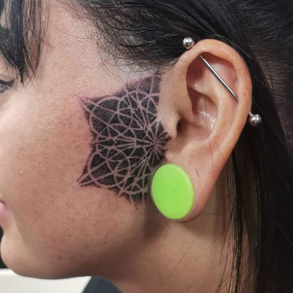 Geometric tribal tattoo on the cheek, featuring a star-like design with intricate linework and shading extending towards the ear