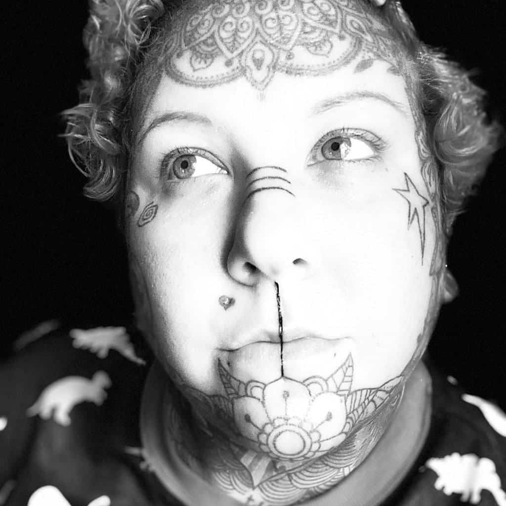 A woman with intricate facial and forehead tattoos, including a mandala design, heart, and stars