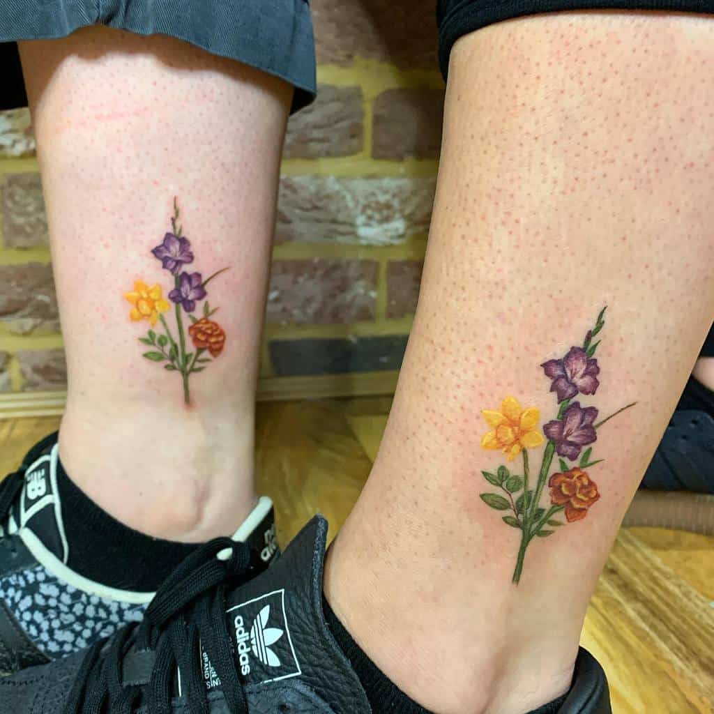 Two legs with matching flower tattoos, featuring purple, yellow, and orange gladiolus blossoms near the ankles