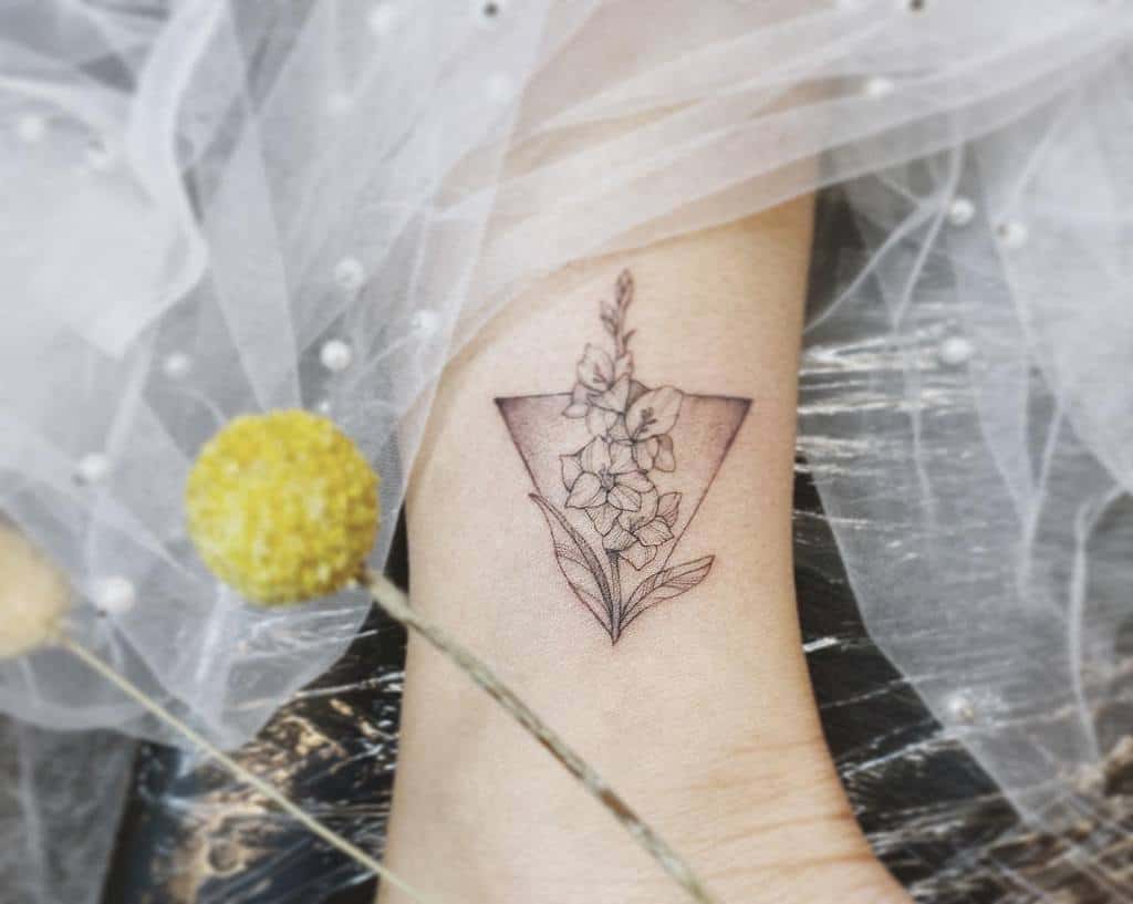 Tattoo of a gladiolus flower within a geometric shape on an ankle, surrounded by white fabric and yellow flowers