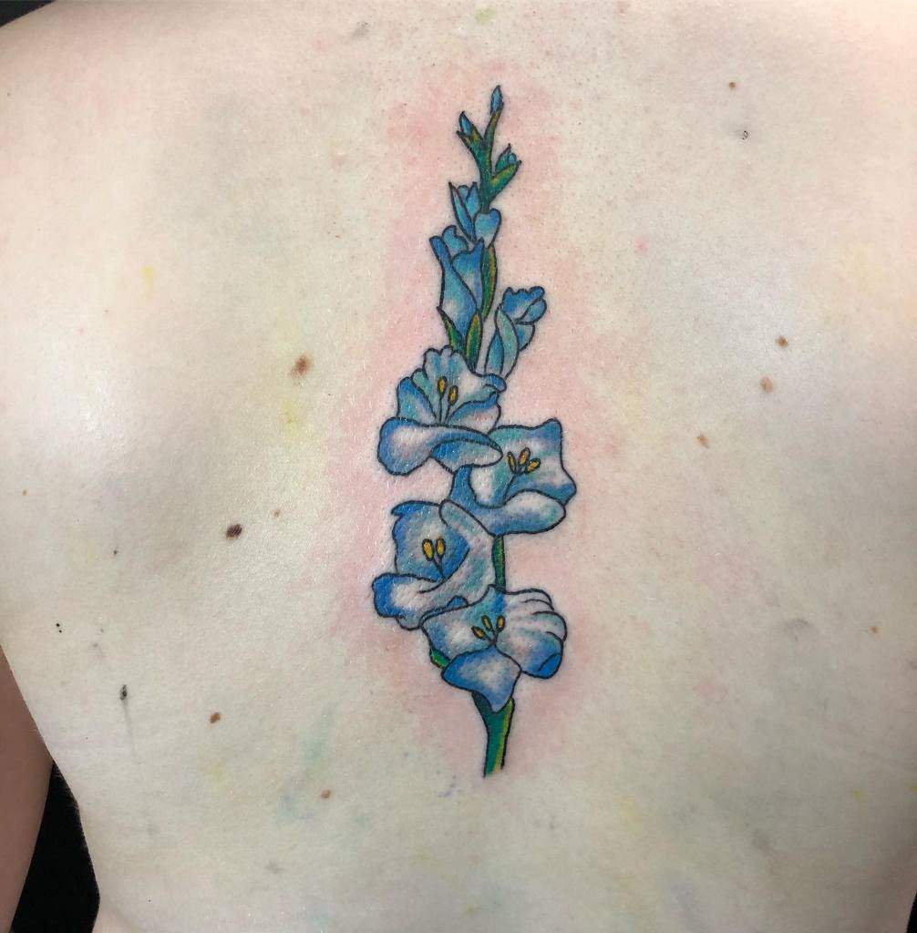 A tattoo of a vertical blue gladiolus with a green stem and yellow centers adorns a person's back