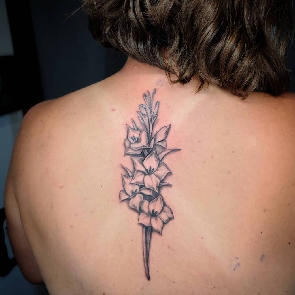Tattoo of flowers, including a single gladiolus stem, on a person's upper back with wavy hair visible