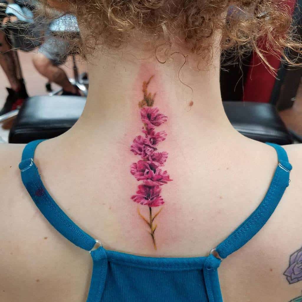 A back view of a person with curly hair showcasing a vertical pink gladiolus tattoo on the neck
