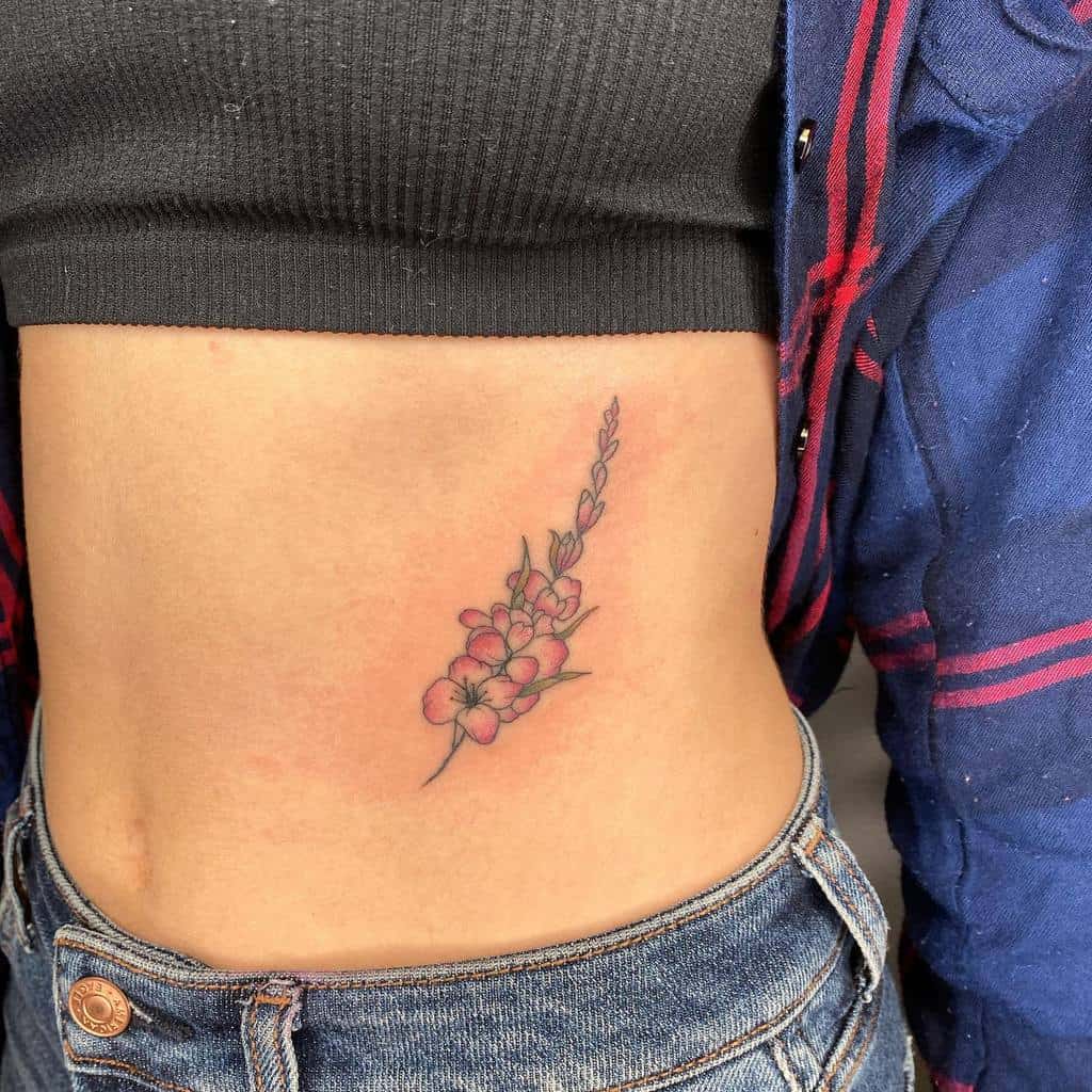 A side tattoo of pink gladiolus flowers and green leaves graces a person's torso, peeking out from under a black crop top