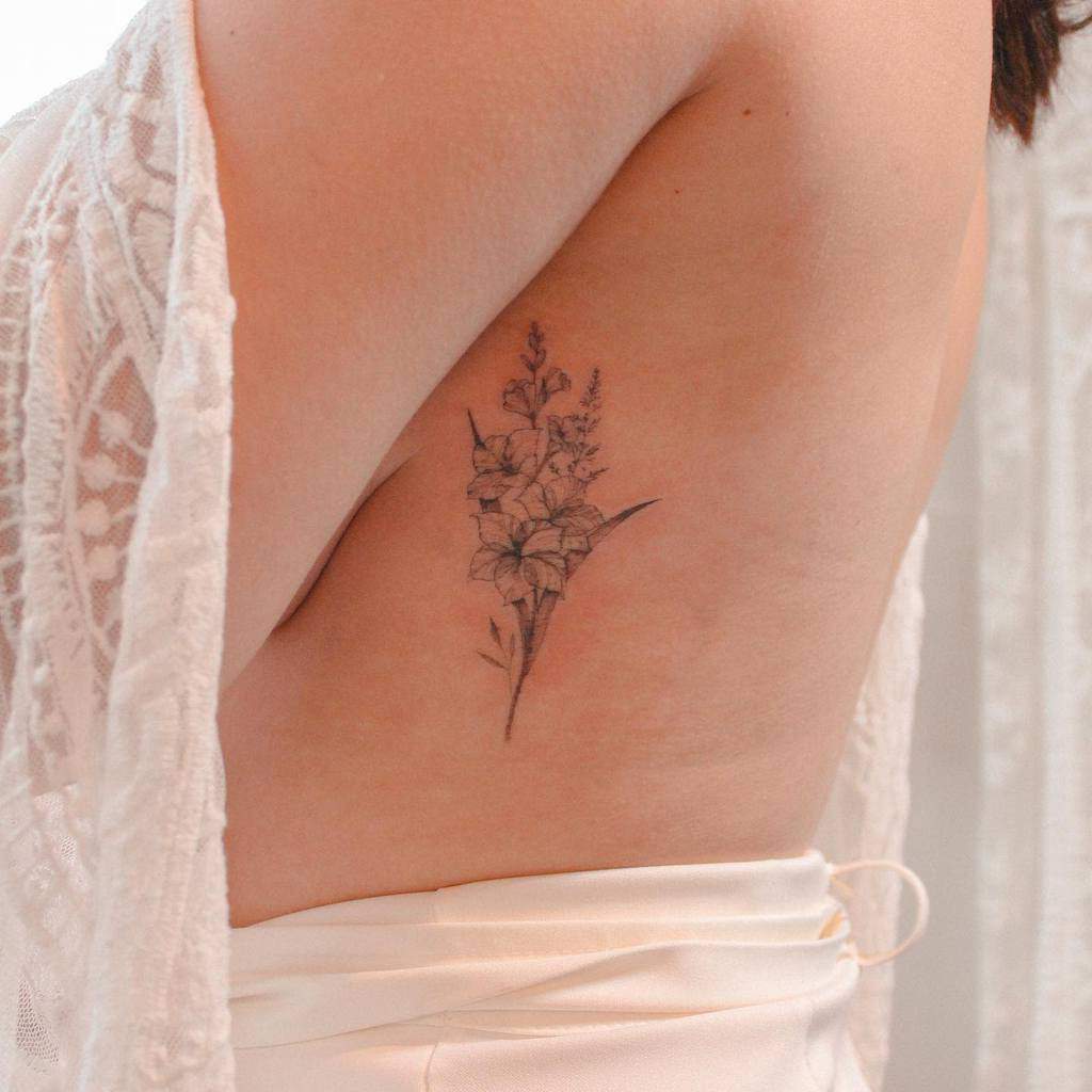 A detailed floral tattoo on a person's ribcage, partially covered by a lace garment, features an intricate gladiolus bloom