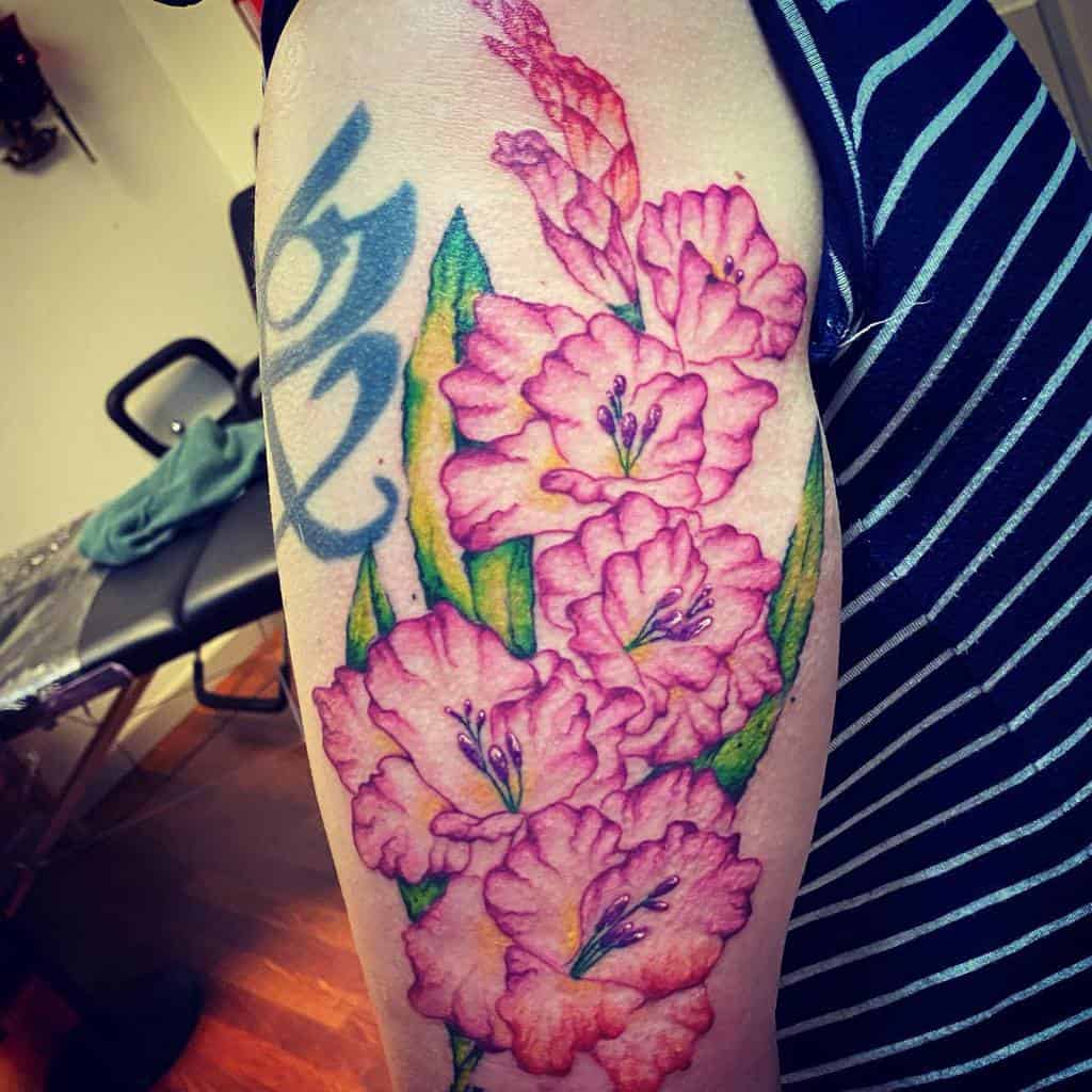 A colorful tattoo of pink gladiolus flowers with green leaves on an arm, complemented by a gray symbol above