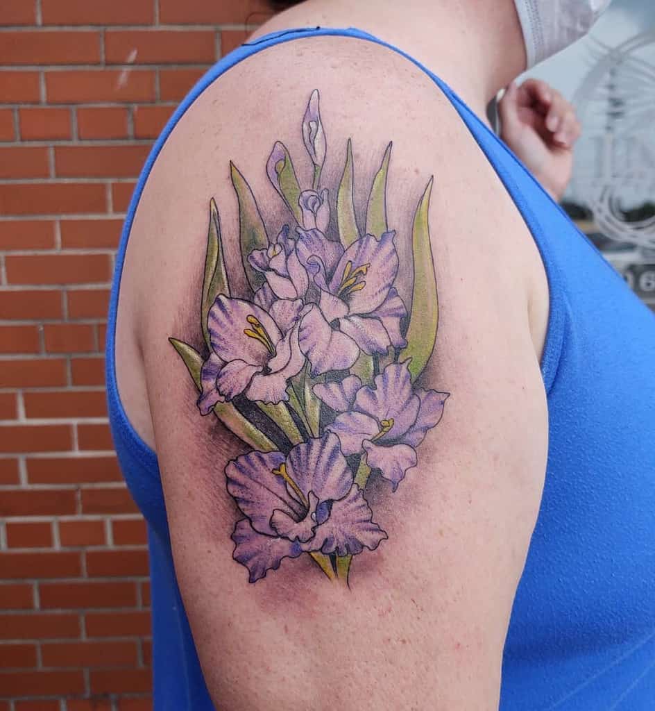 Tattoo of purple gladiolus flowers and green leaves on an upper arm wearing a blue tank top