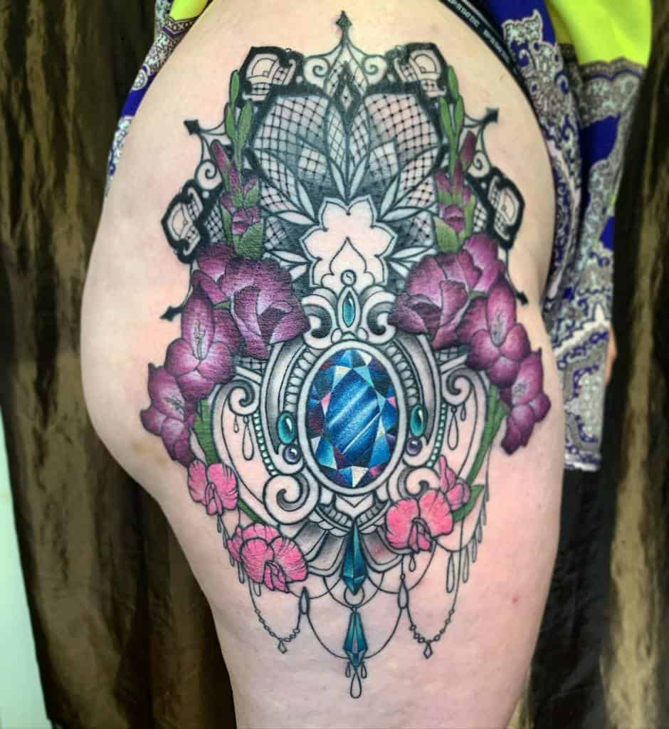 Colorful floral gladiolus tattoo with a blue jewel centerpiece and intricate lace detailing on a thigh