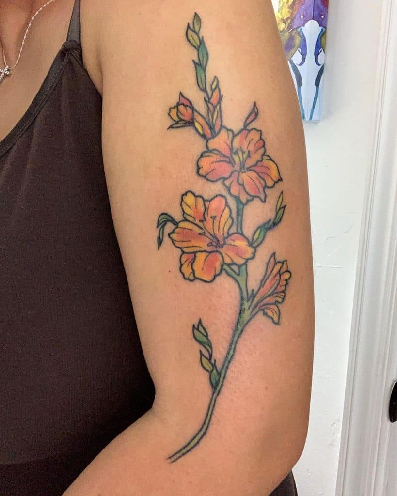 A tattoo of gladiolus flowers, with their vibrant orange petals and lush green leaves, graces an upper arm