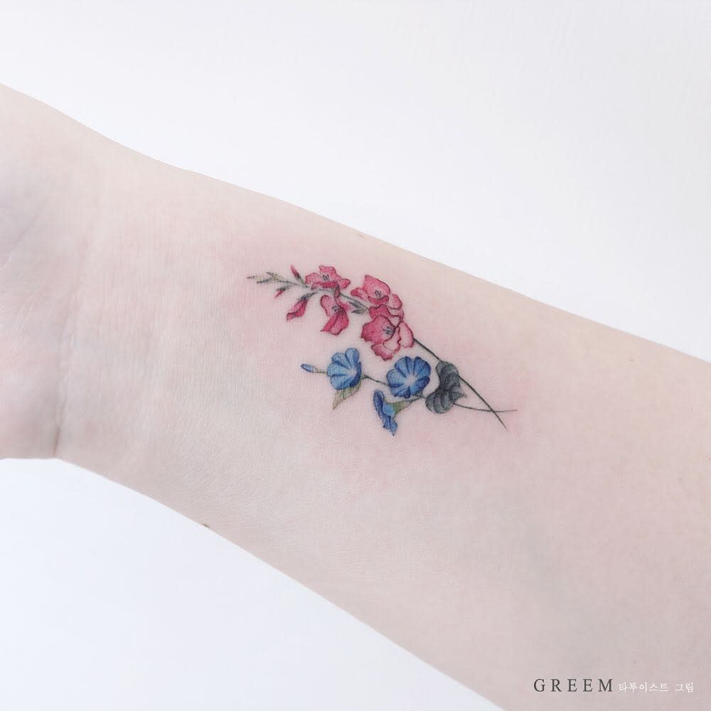 A delicate tattoo of pink gladiolus and blue flowers graces a person's inner forearm
