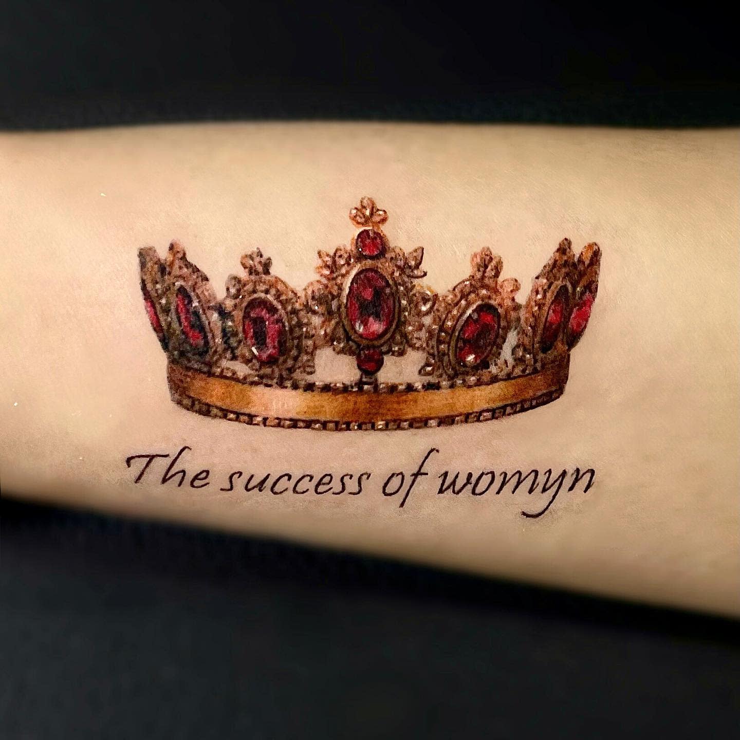 Tattoo of a jeweled crown above the text "The success of womyn" on an arm