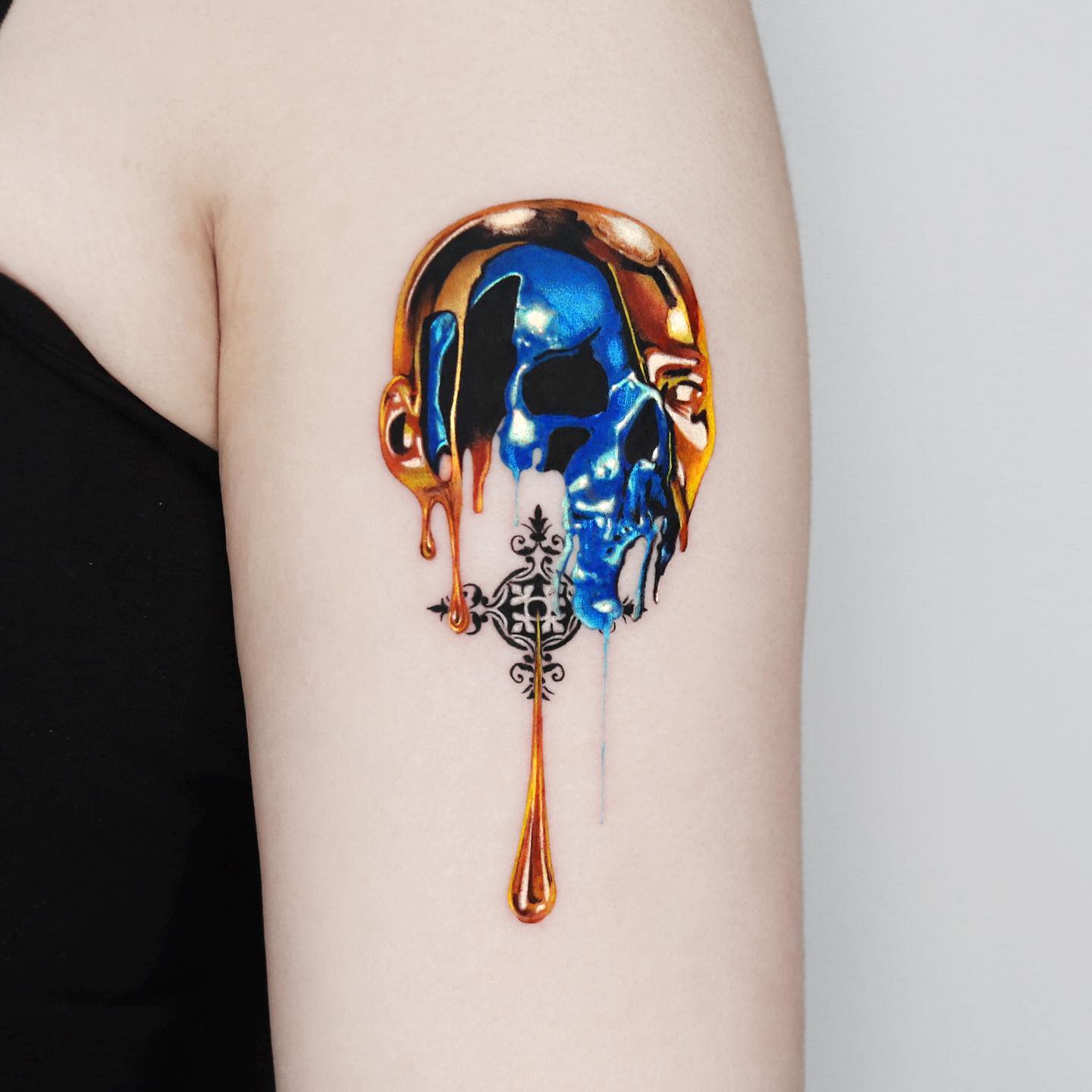 A colorful melting skull tattoo on an upper arm with an intricate design beneath it