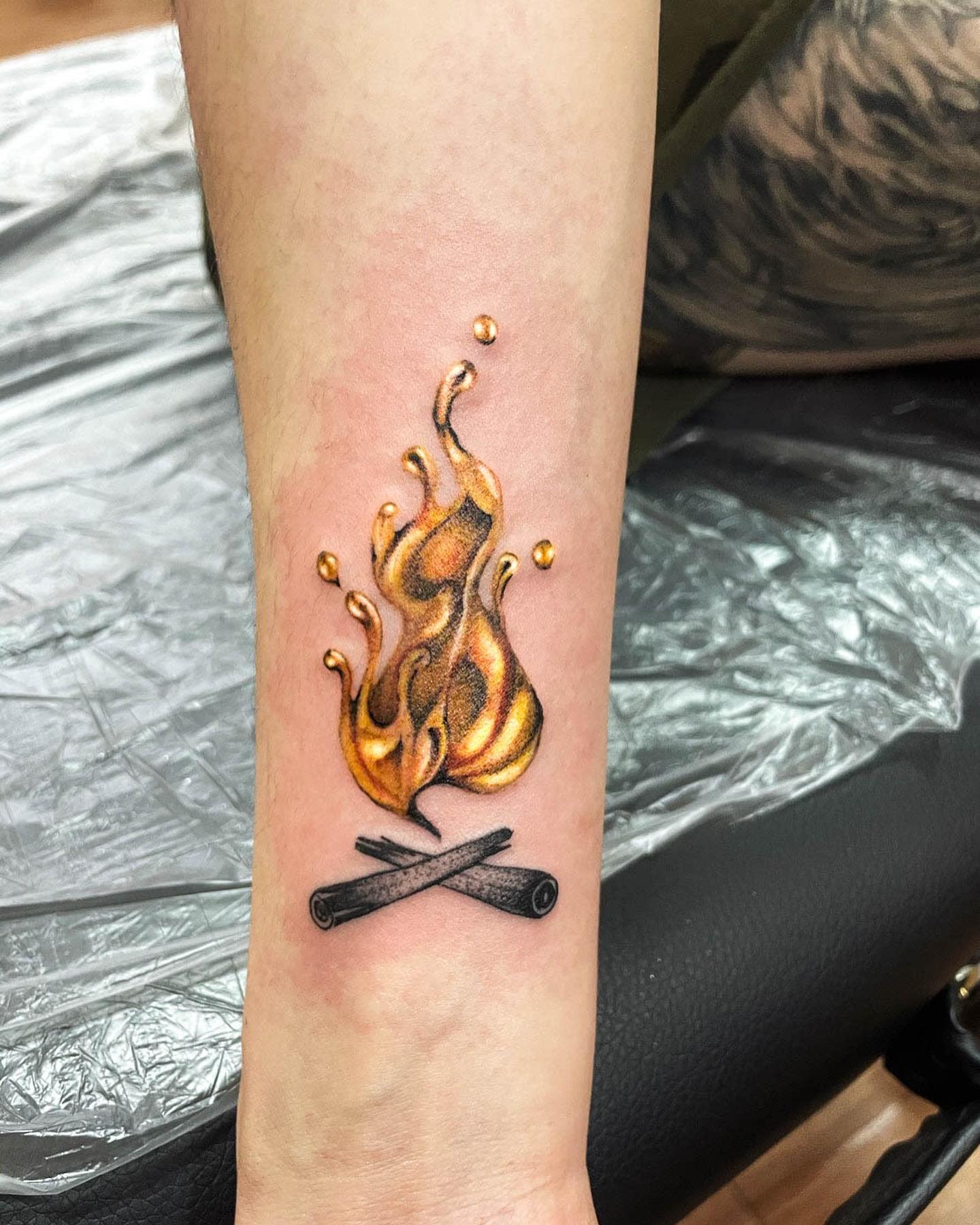 A realistic tattoo of a golden flame and crossed sticks on an inner forearm