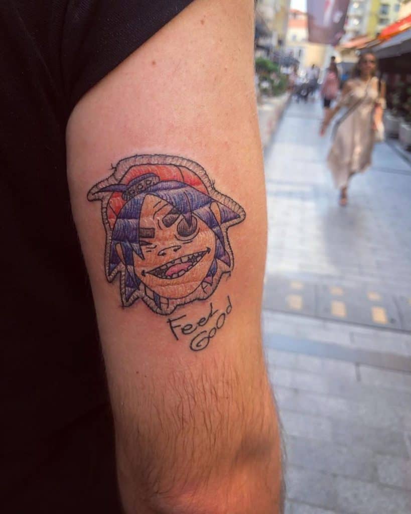 Tattoo of a grinning animated character with an embroidered texture, featuring detailed stitching and a playful handwritten phrase.