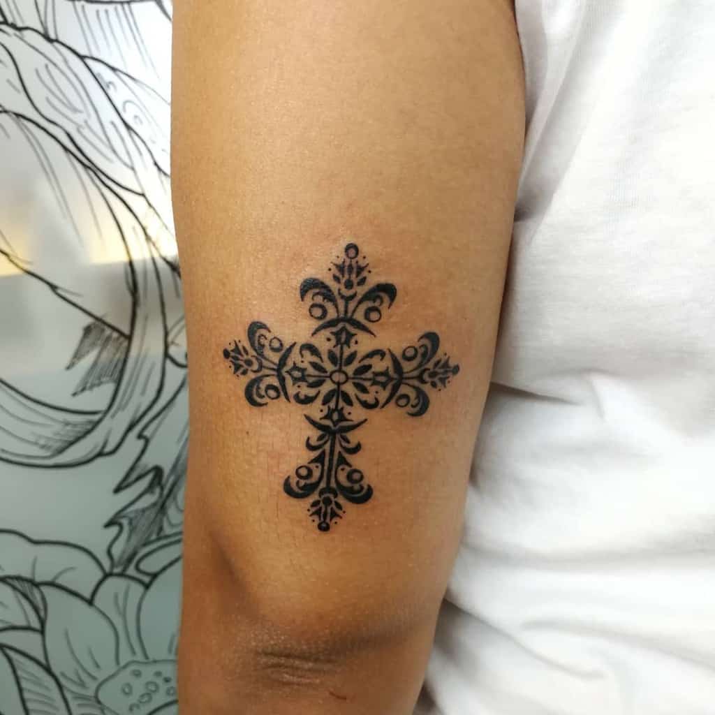 Intricate black cross tattoo with ornate patterns graces the upper arm