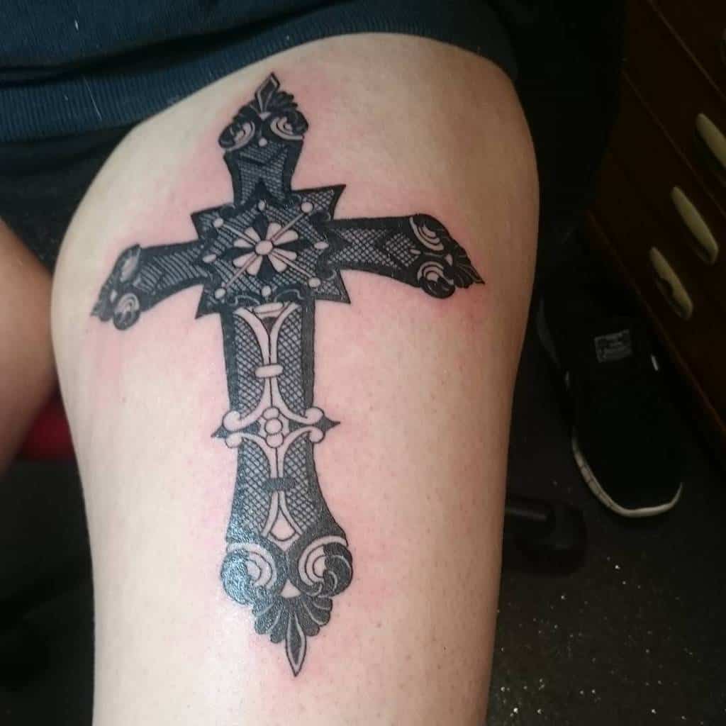 This delicate tattoo on a person's thigh boasts intricate black lace-style patterns, seamlessly blending floral and geometric designs with the cross at its center