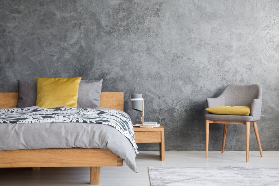 Minimalist bedroom with a wooden bed, gray and yellow pillows, a small bedside table with a lamp, and a gray upholstered chair against a concrete wall