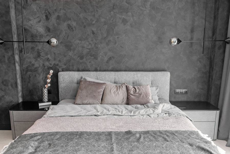 Modern bedroom with a gray textured wall, a gray headboard, neatly arranged pillows, and matching side tables with wall-mounted lamps