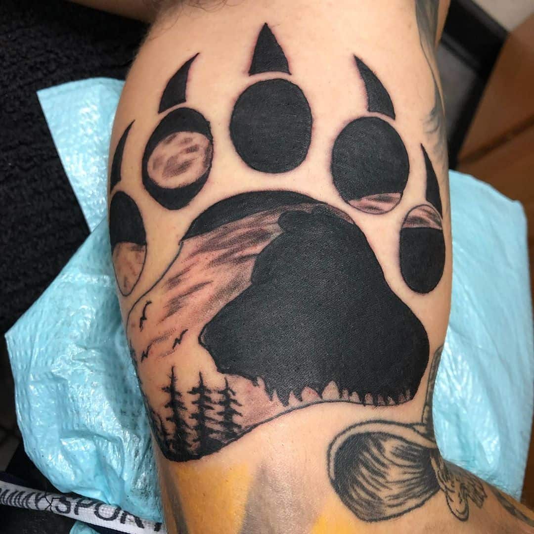 Arm tattoo of a bear paw featuring an intricate nature scene with moon phases and trees seamlessly woven into the design