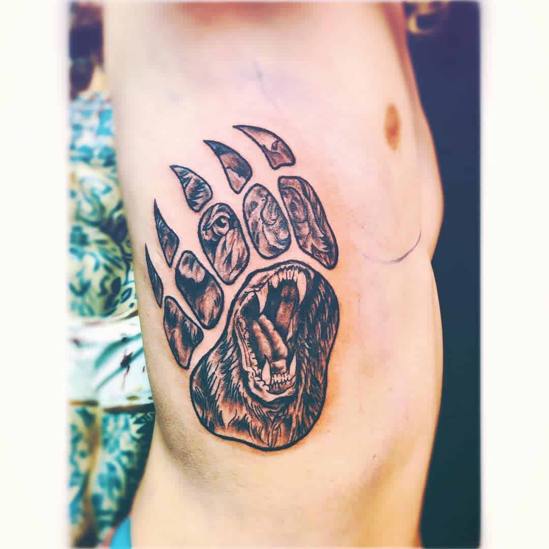 Tattoo of a bear's open mouth integrated within a detailed bear paw design on the side of a person's torso