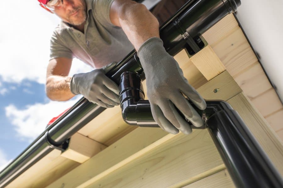 Person wearing gloves installs a black rain gutter on the edge of a wooden roof under a blue sky