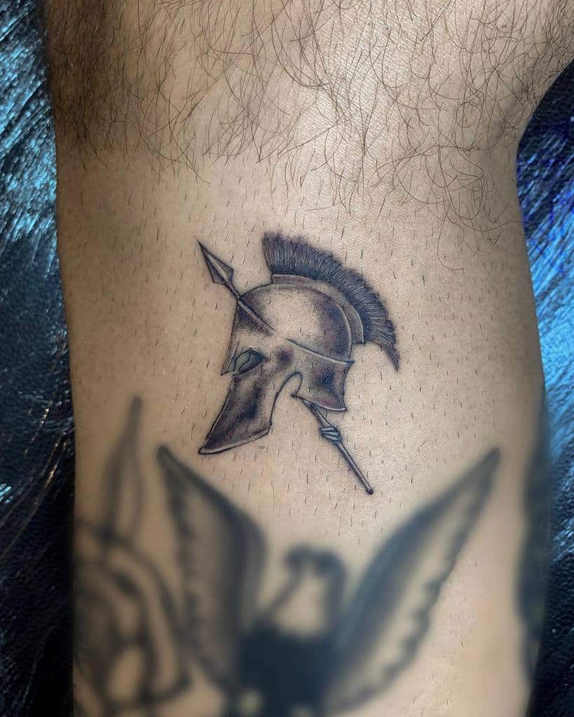 Tattoo of a Spartan helmet with an arrow through it, rendered with fine single needle detail on a forearm