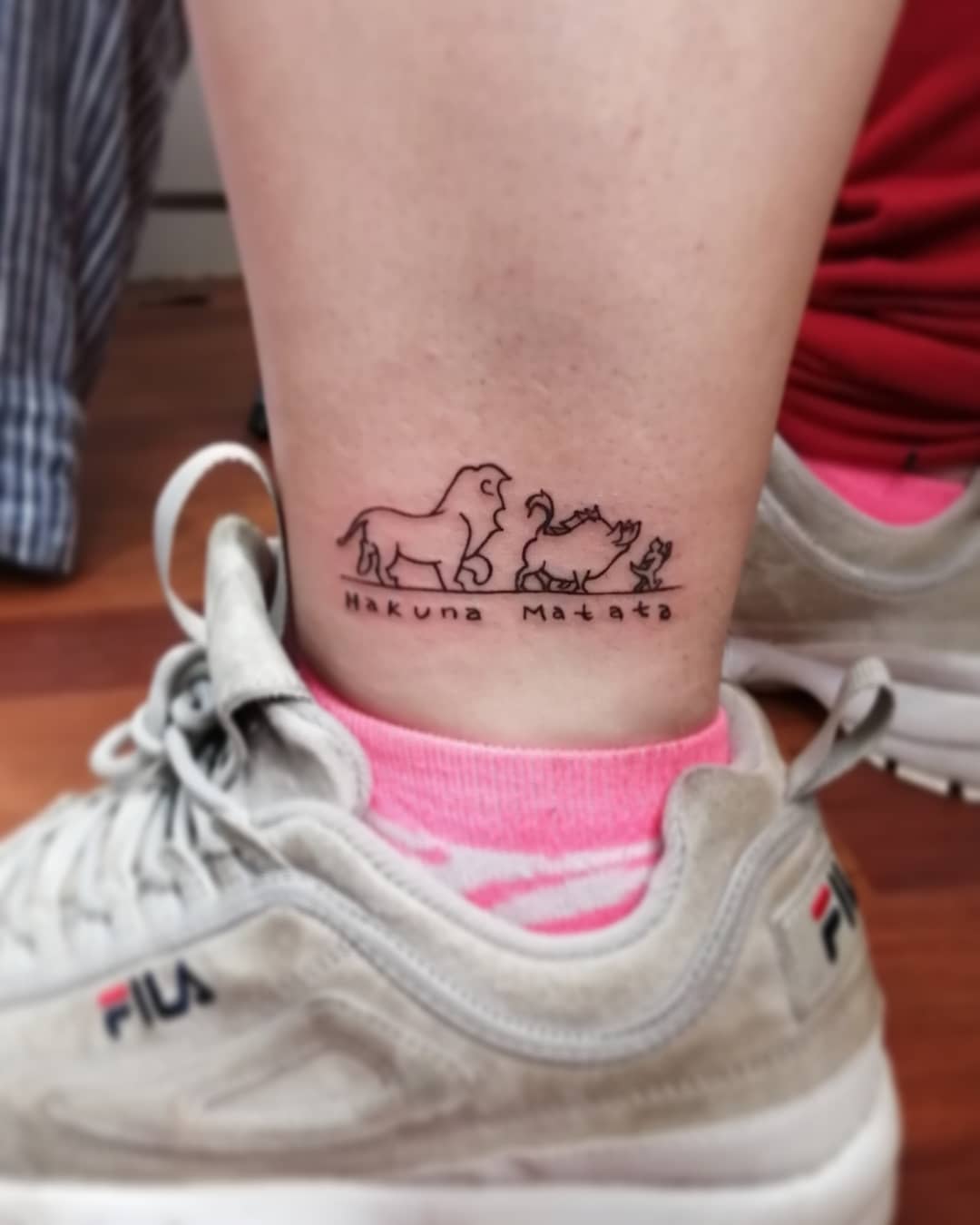 Tattoo of a lion, warthog, and meerkat with "Hakuna Matata" on someone's ankle.