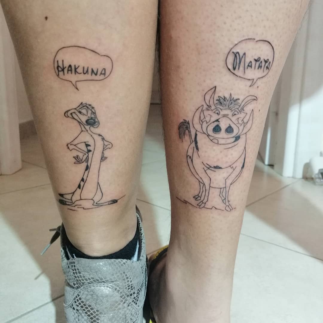 Two legs with matching tattoos of Timon and Pumbaa saying "Hakuna Matata"