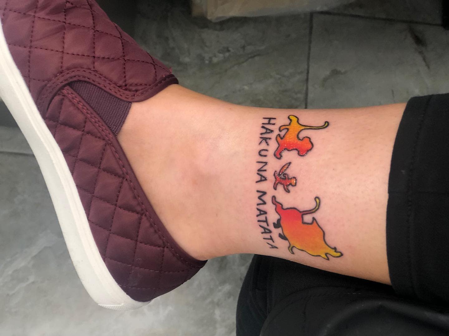 An ankle tattoo with silhouettes of Timon, Pumbaa, and Simba, and the text "Hakuna Matata"