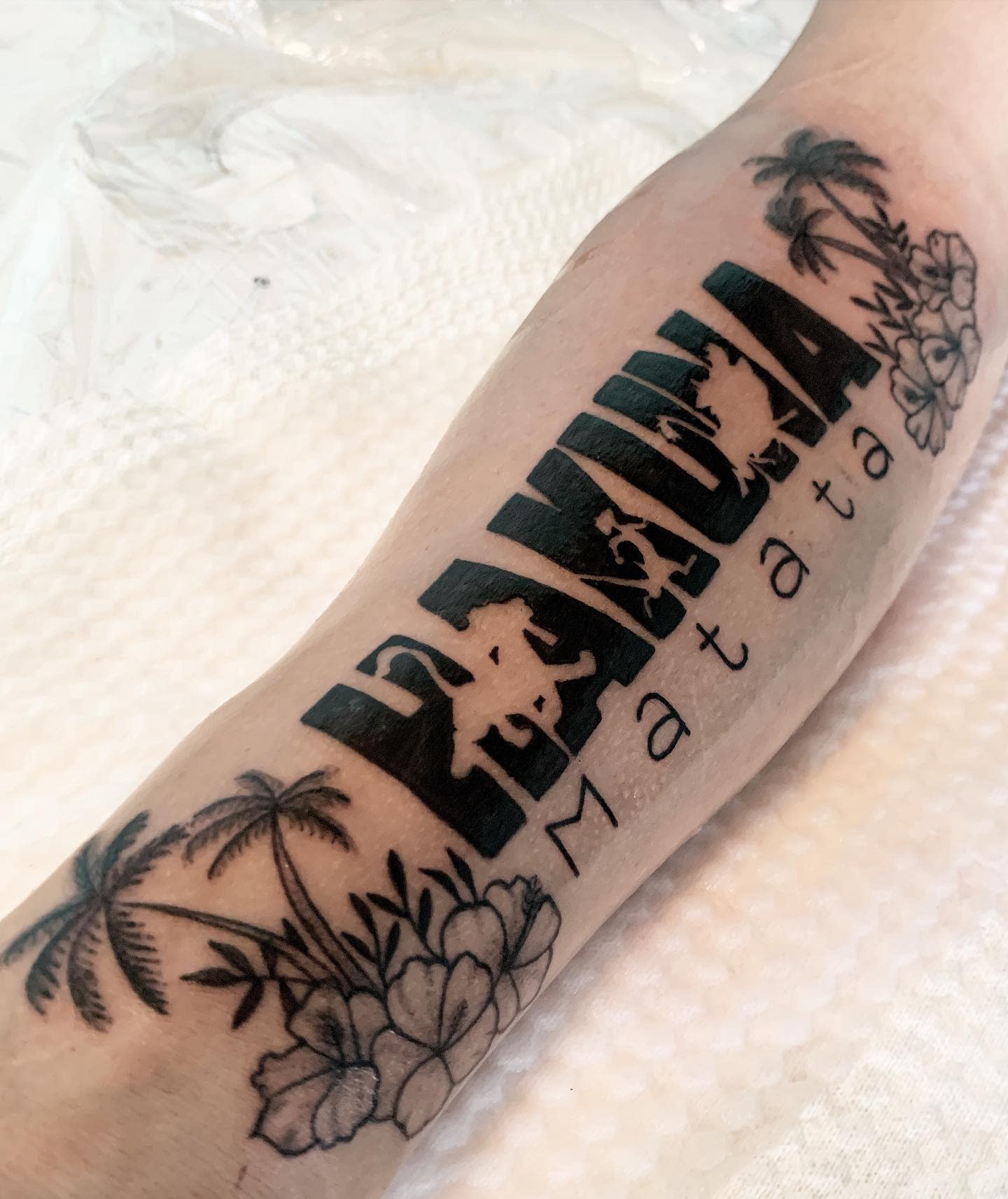 Forearm tattoo with "Hakuna Matata" text, palm trees, and flowers in black ink