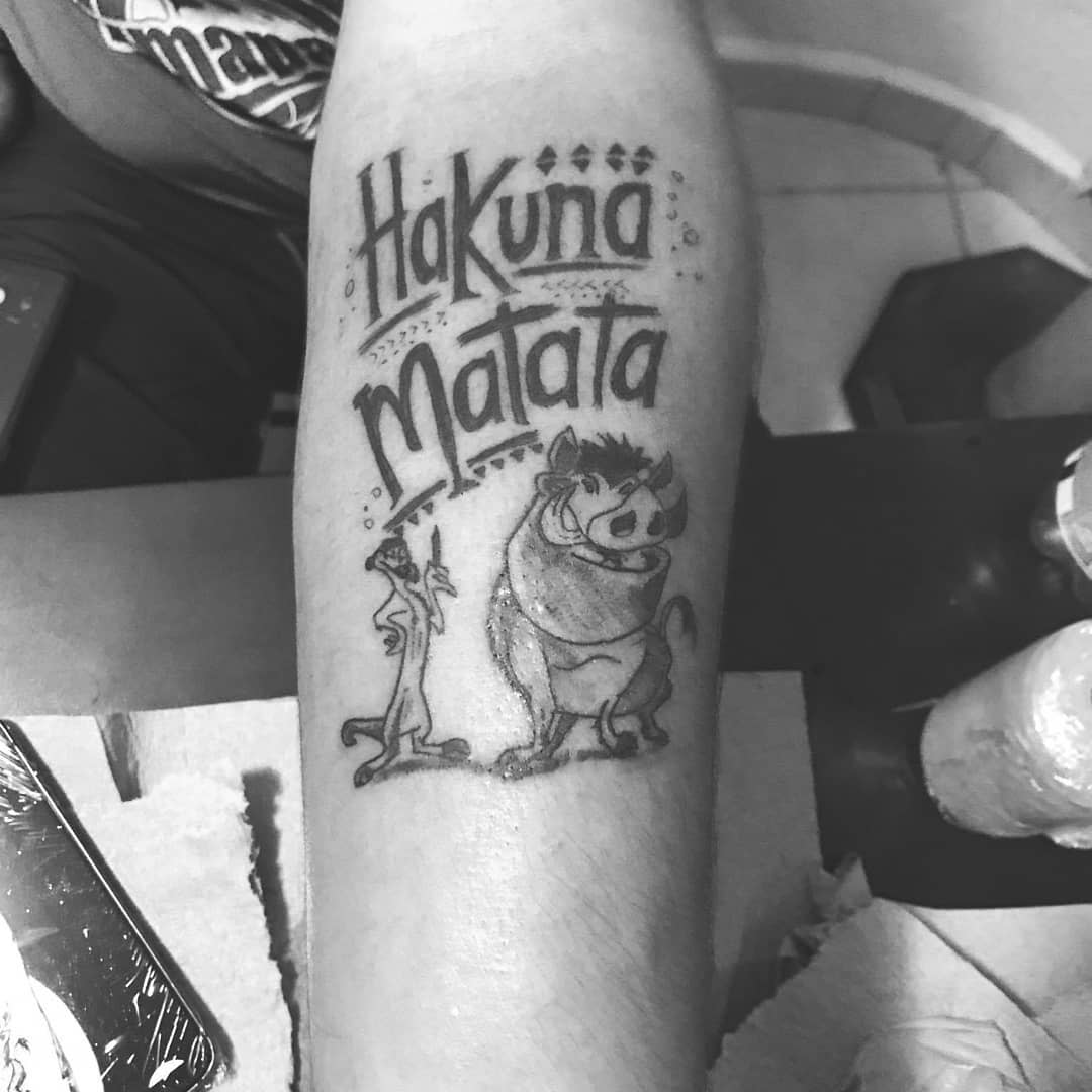 Tattoo of "Hakuna Matata" with cartoon characters in black ink on a person's forearm