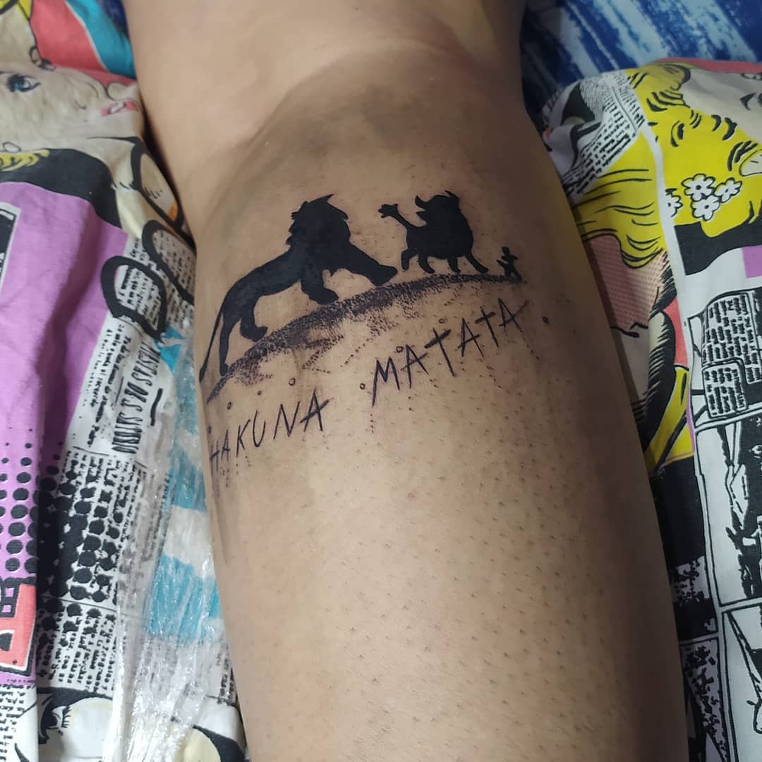 Tattoo of "Hakuna Matata" with silhouettes of a lion, warthog, and meerkat on a leg