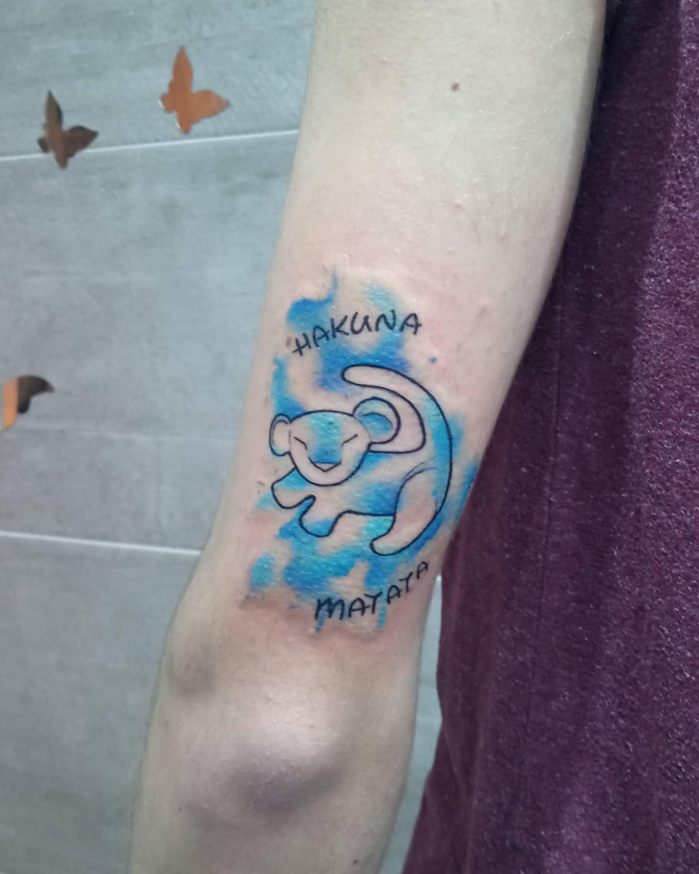 Colorful watercolor Hakuna Matata tattoo featuring a playful lion cub on the upper arm