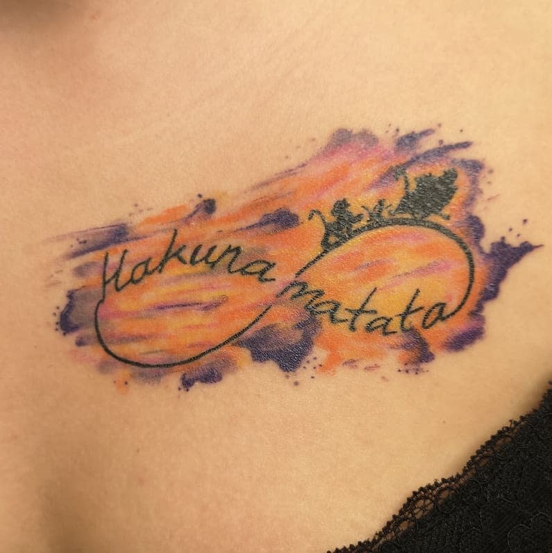 Flowing Hakuna Matata script tattoo with a sunset-colored watercolor background and silhouette