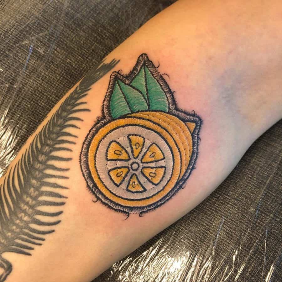 Tattoo of a sliced orange with green leaves, designed in an embroidery style with realistic stitched textures.