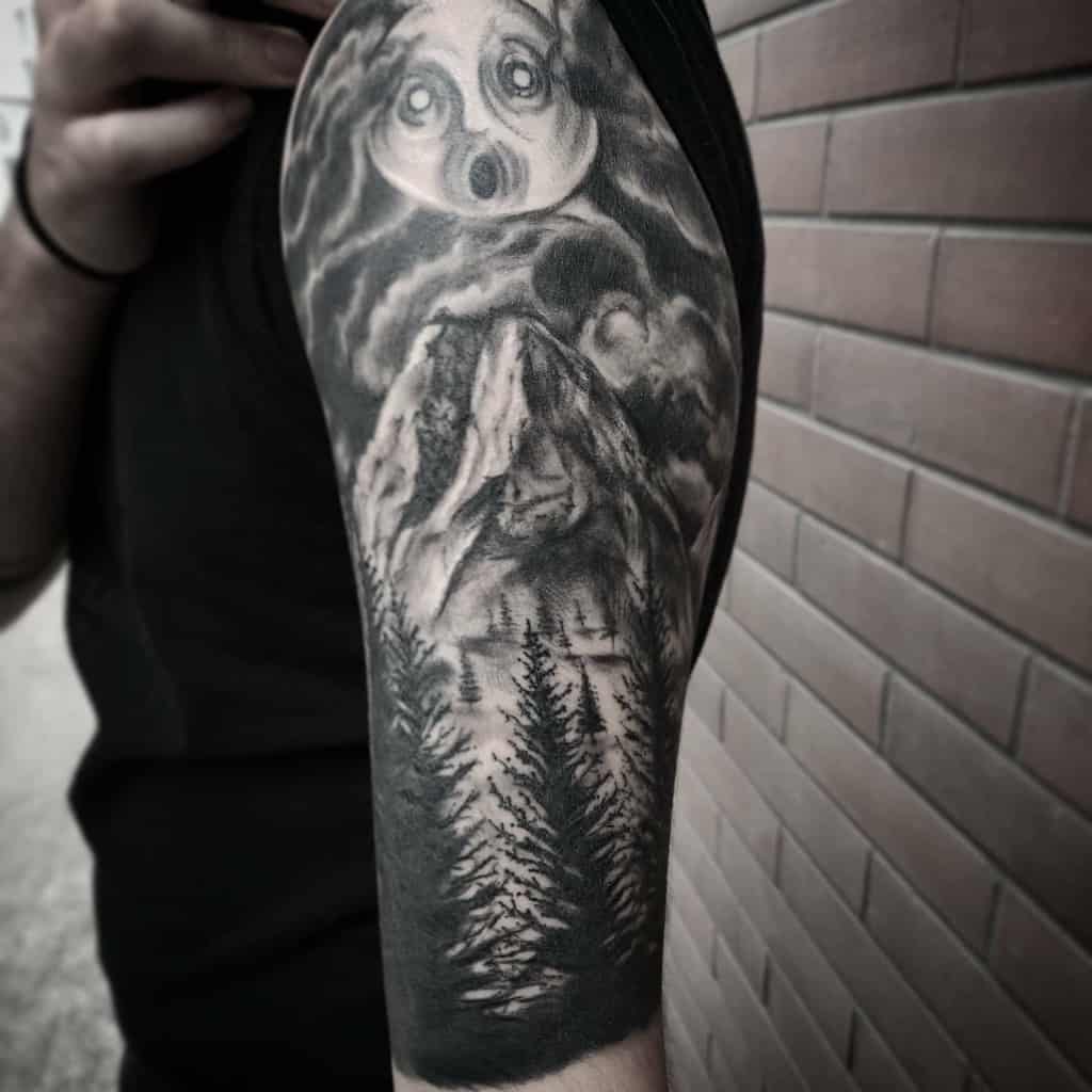 Black-and-grey half-sleeve tattoo depicting a hauntingly surreal moon above towering mountains and dense pine trees, creating a dramatic forest scene