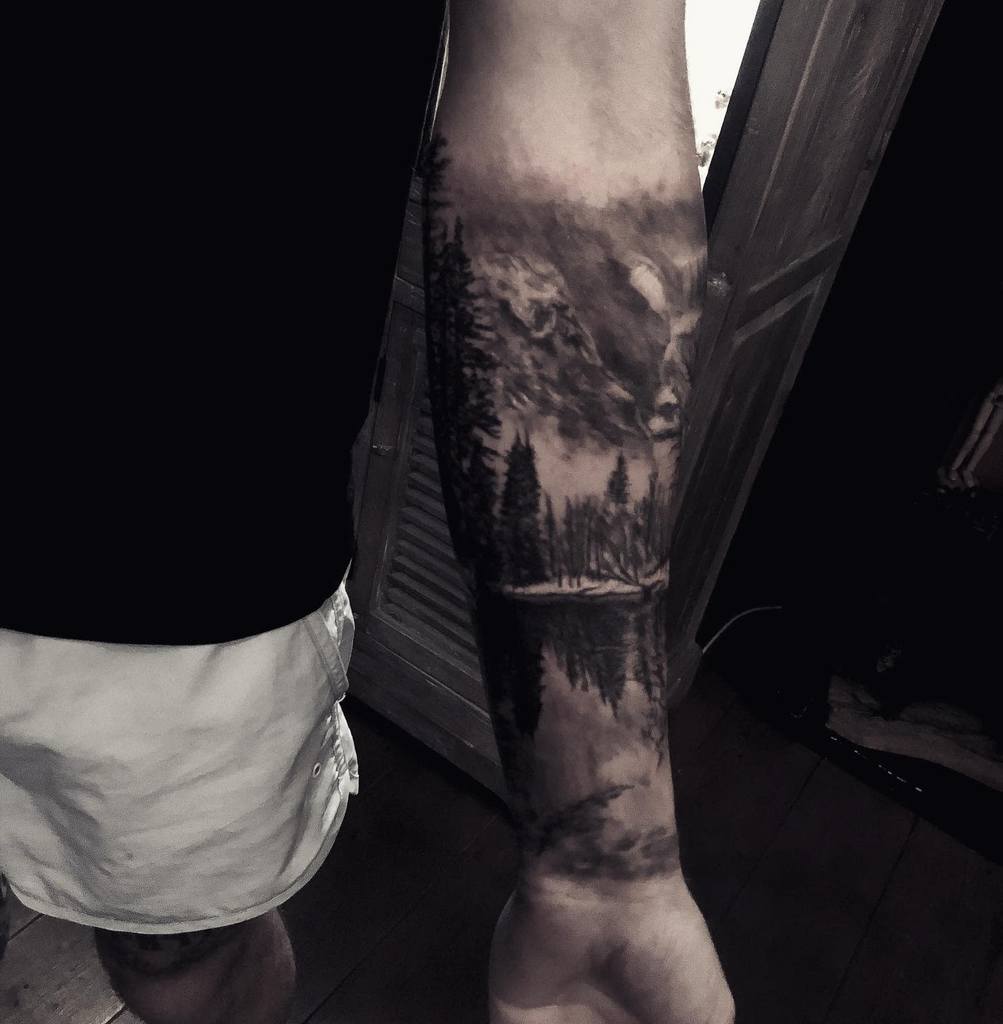 Forearm tattoo featuring a mountain landscape with trees and a reflection in the water