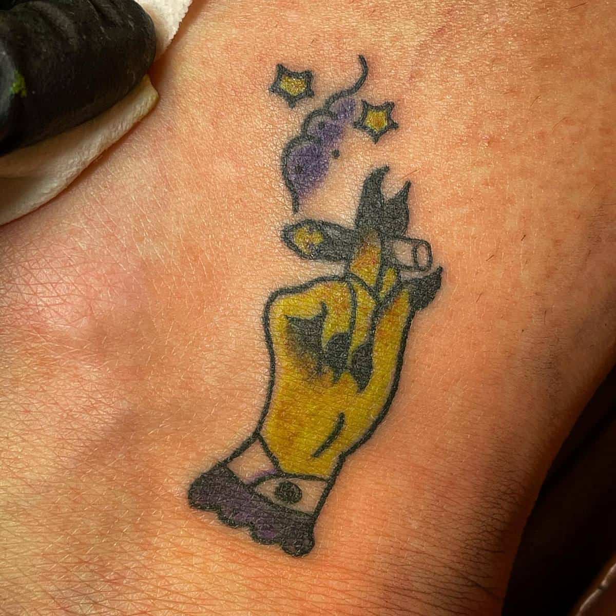 A tattoo of a witch hand holding a lit joint with smoke and stars floating above