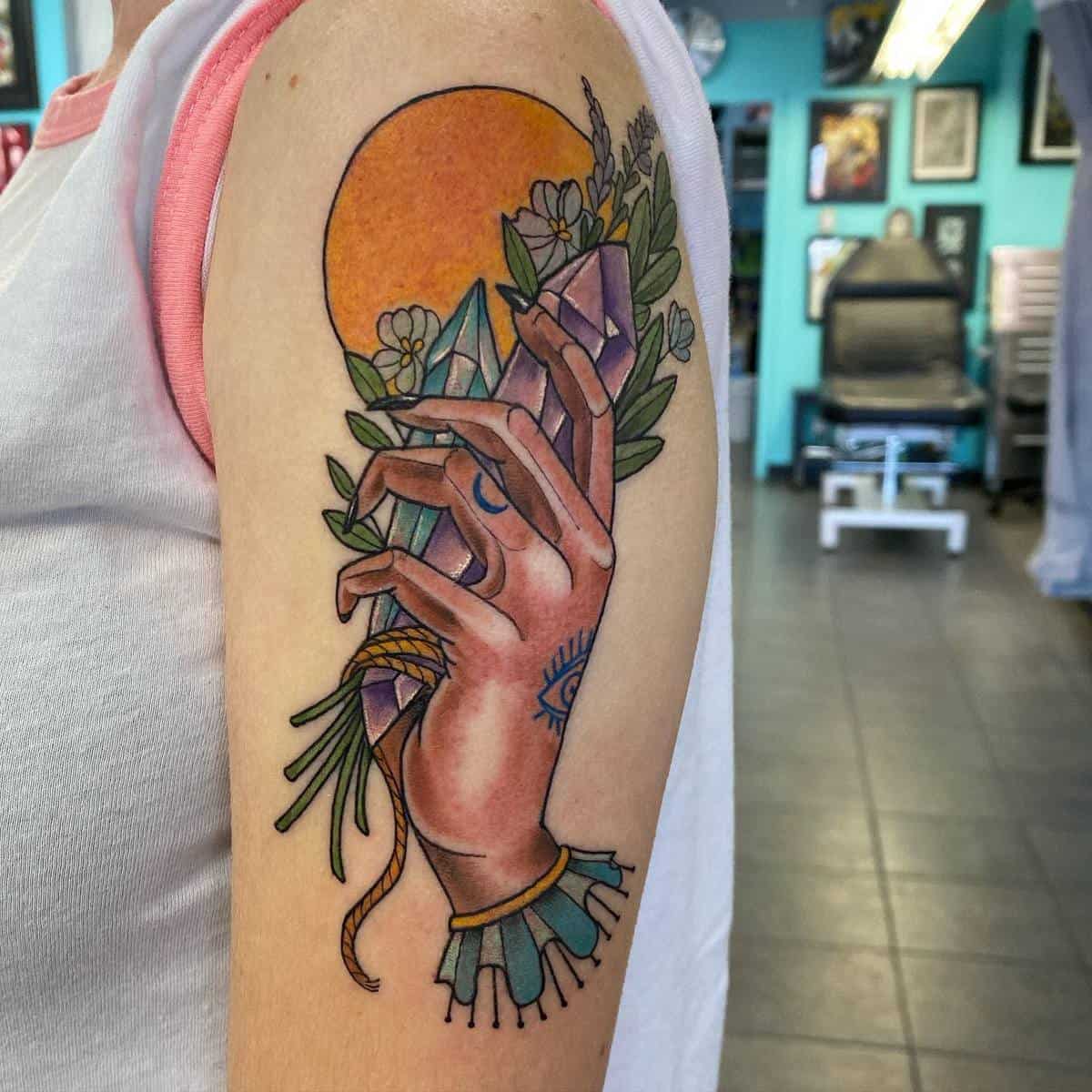 Upper arm tattoo of a hand holding crystals, with flowers and a sun in the background