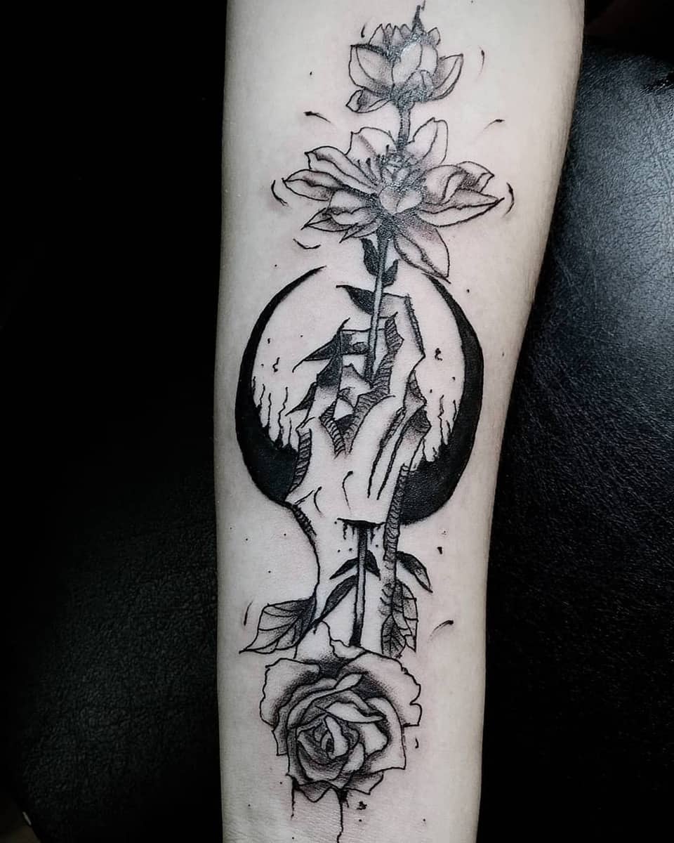 Hand holding a blooming flower with a crescent moon tattoo on forearm