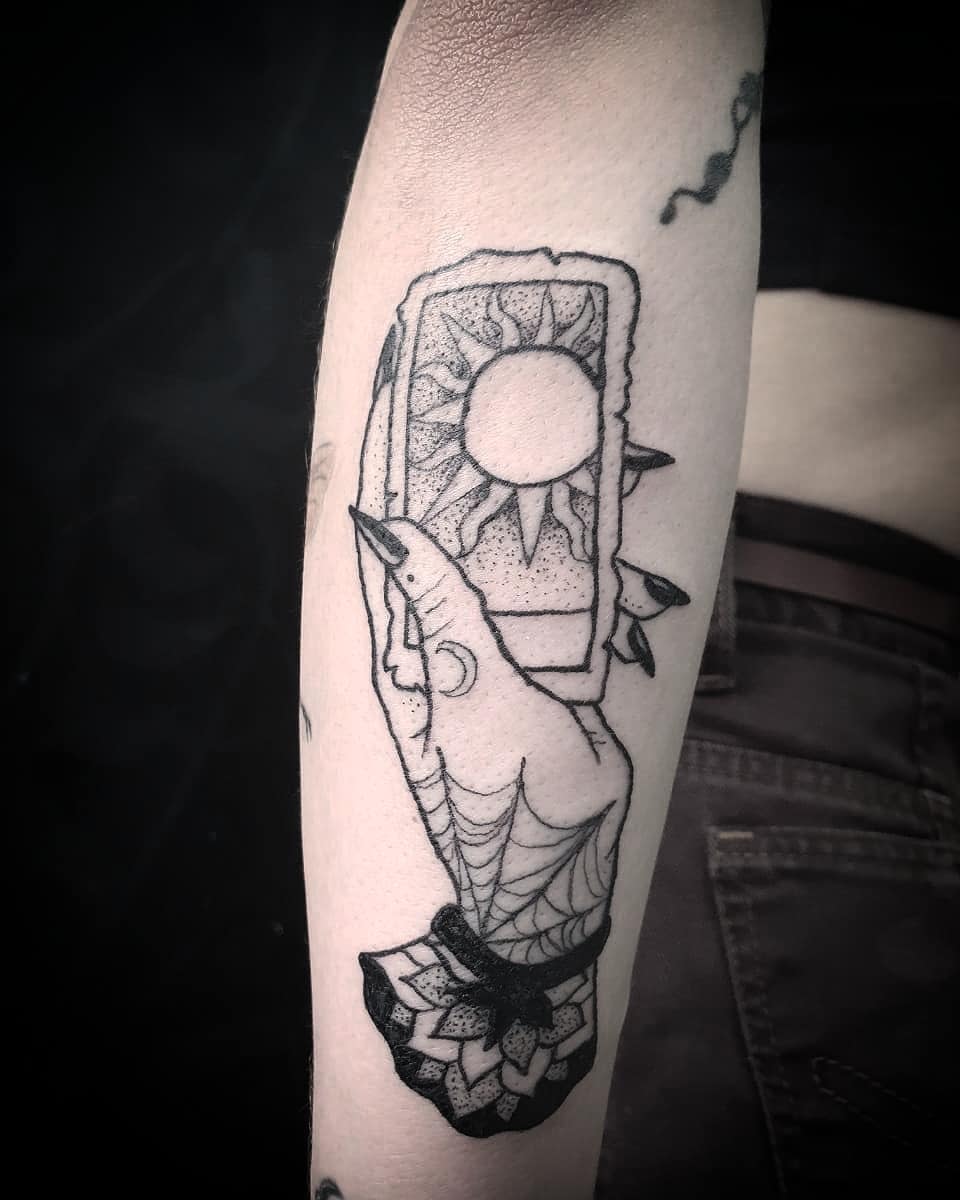 Hand holding a sun tarot card with spiderweb design tattoo on forearm, symbolizing clarity and mysticism