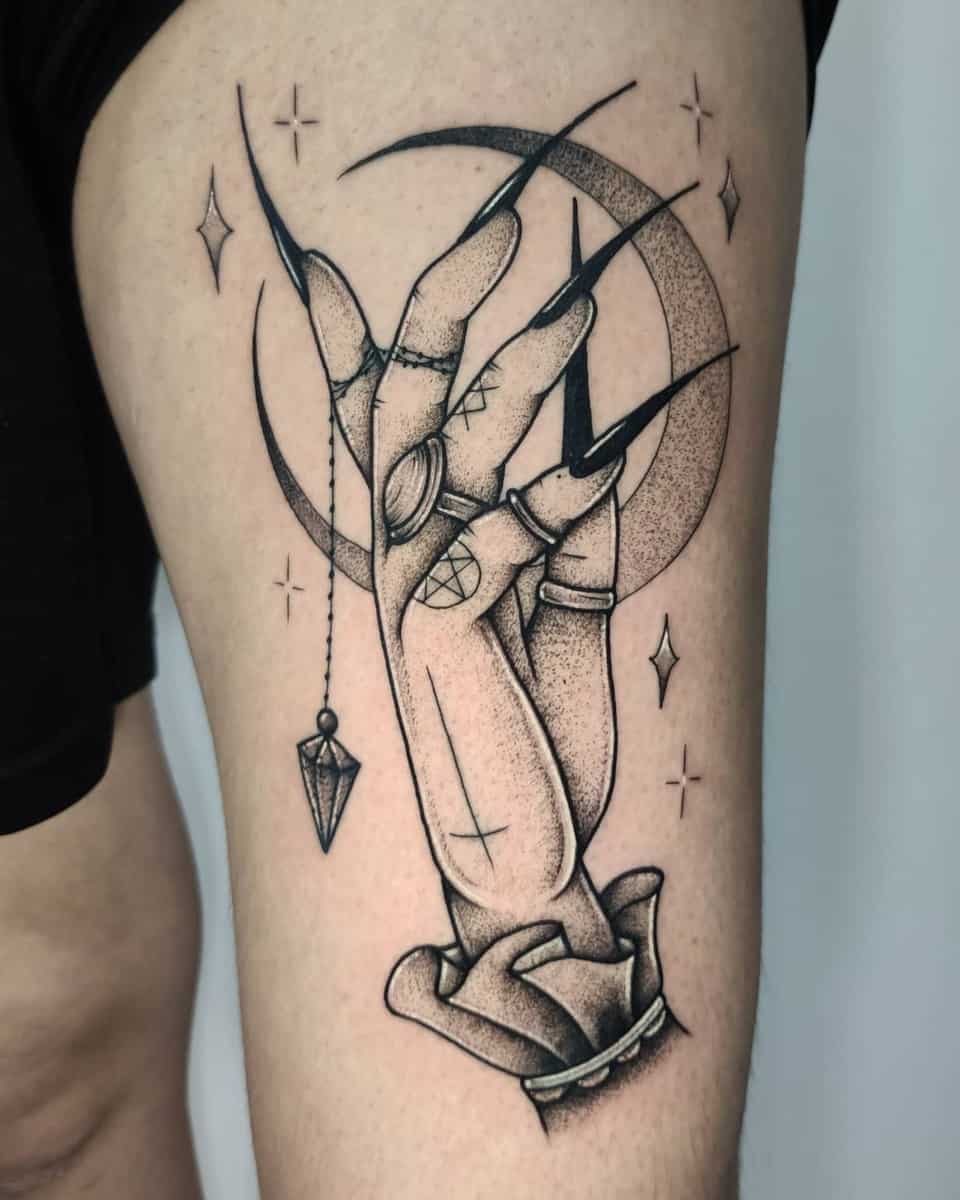 Tattoo of a hand with long nails holding a crystal pendant with stars and a crescent moon