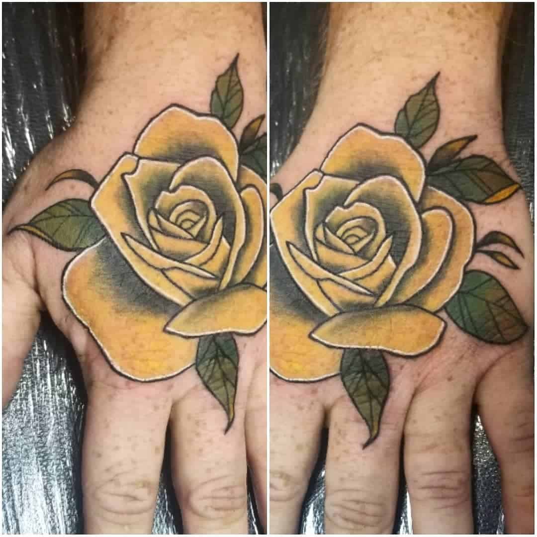 Hand tattoo featuring a yellow and black rose design with green leaves, positioned on the back of the hand