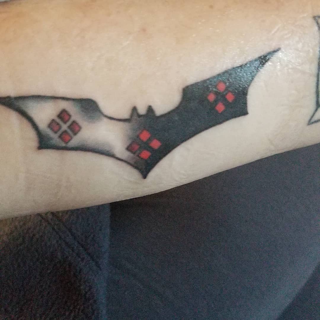 Tattoo of a Batman logo with Harley Quinn's signature diamond patterns inside on the forearm