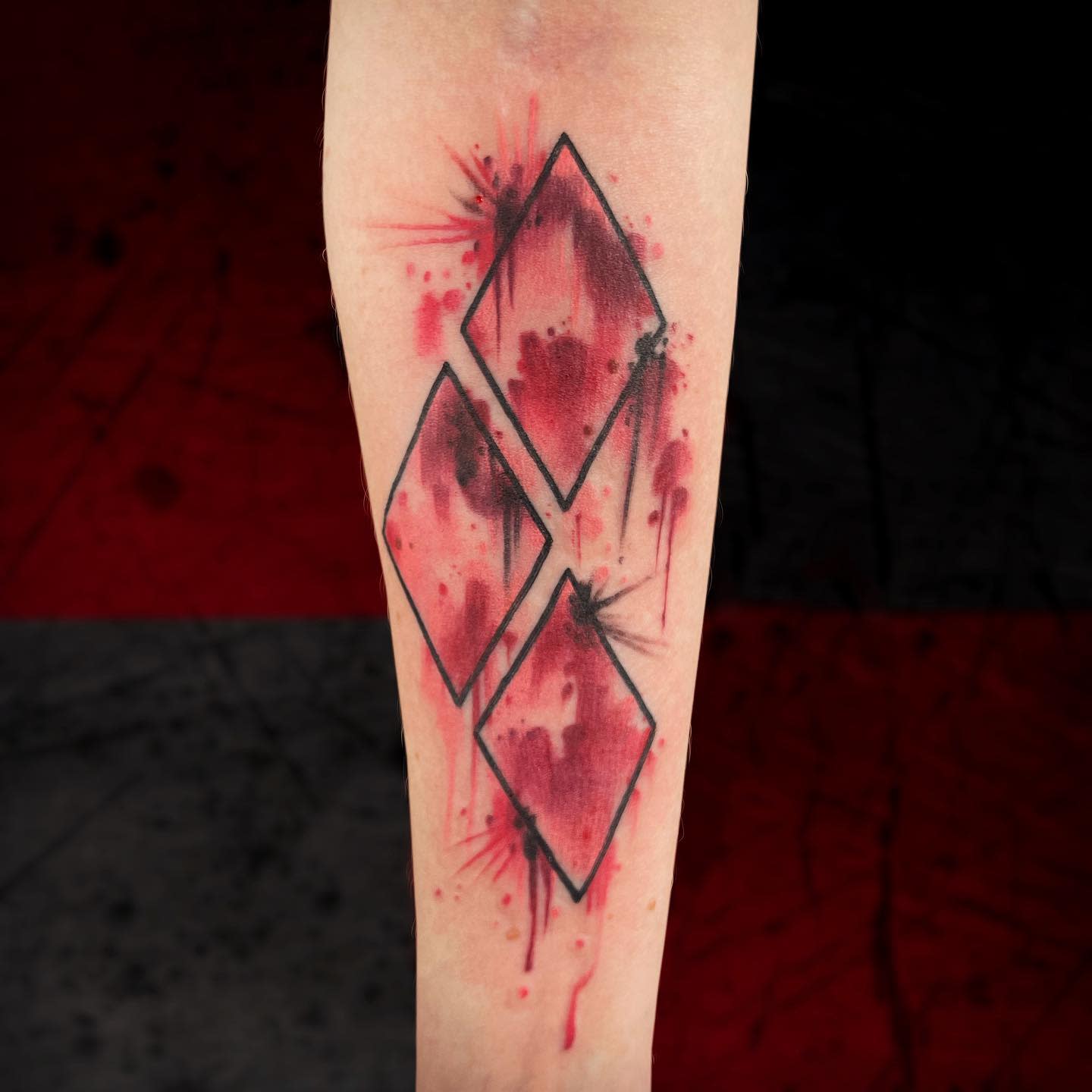 Tattoo featuring three rhombuses with a red splatter background on the inner forearm