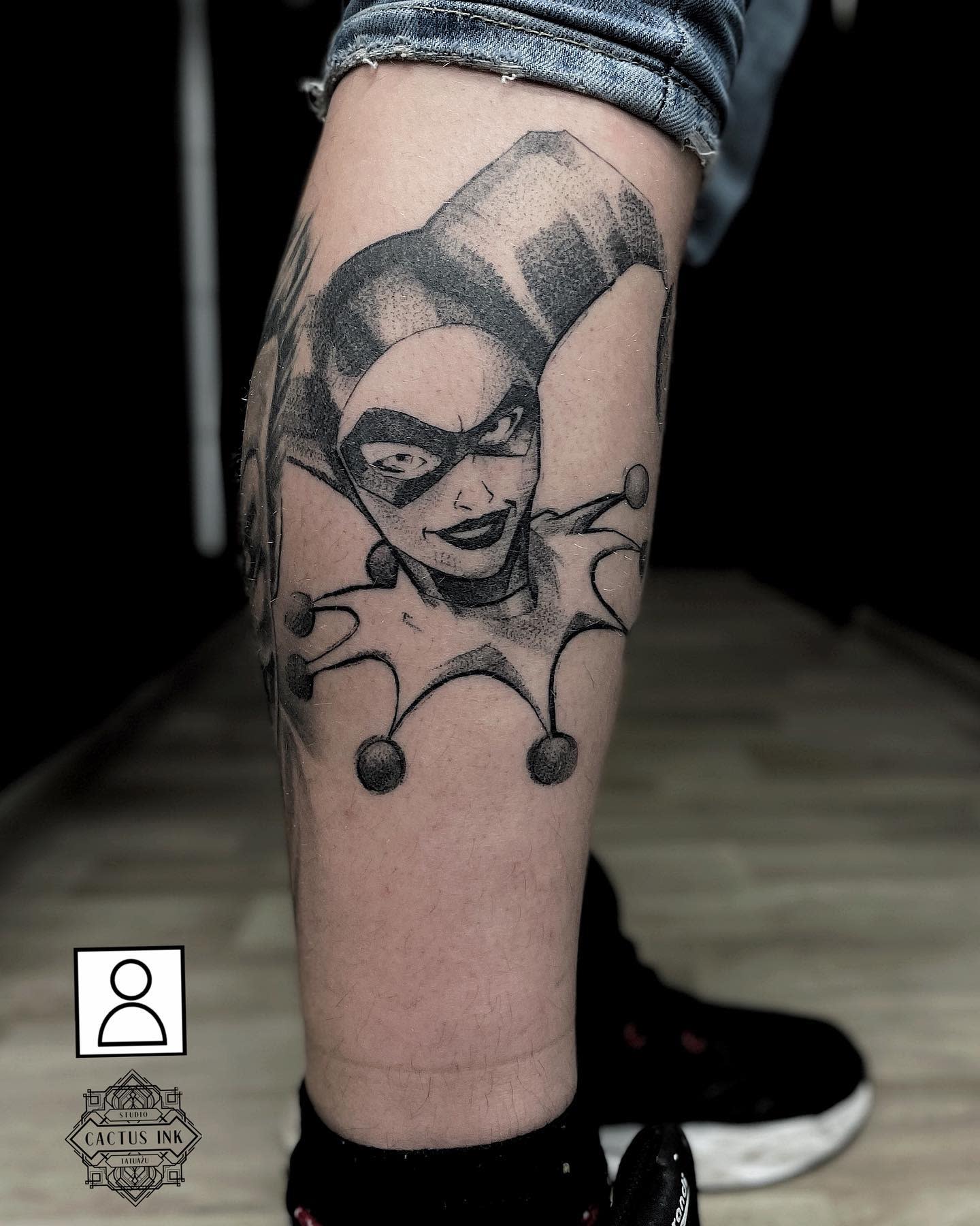 Black and grey tattoo of Harley Quinn's face in her jester costume on the lower leg
