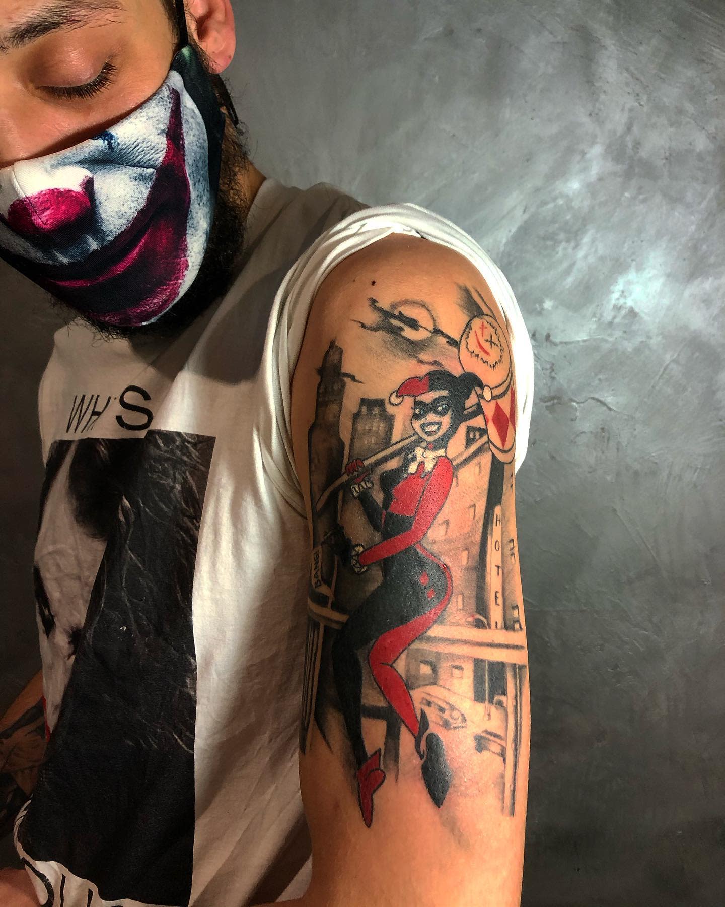 Tattoo of Harley Quinn in her jester outfit holding a mallet, set against a cityscape background on the upper arm