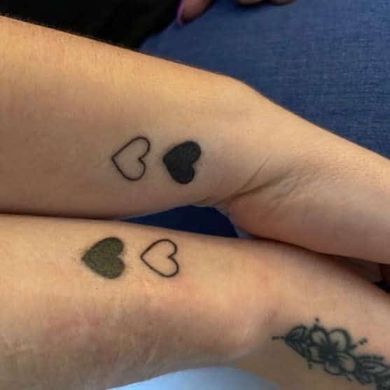 Matching tattoos of black hearts and heart outlines on two forearms
