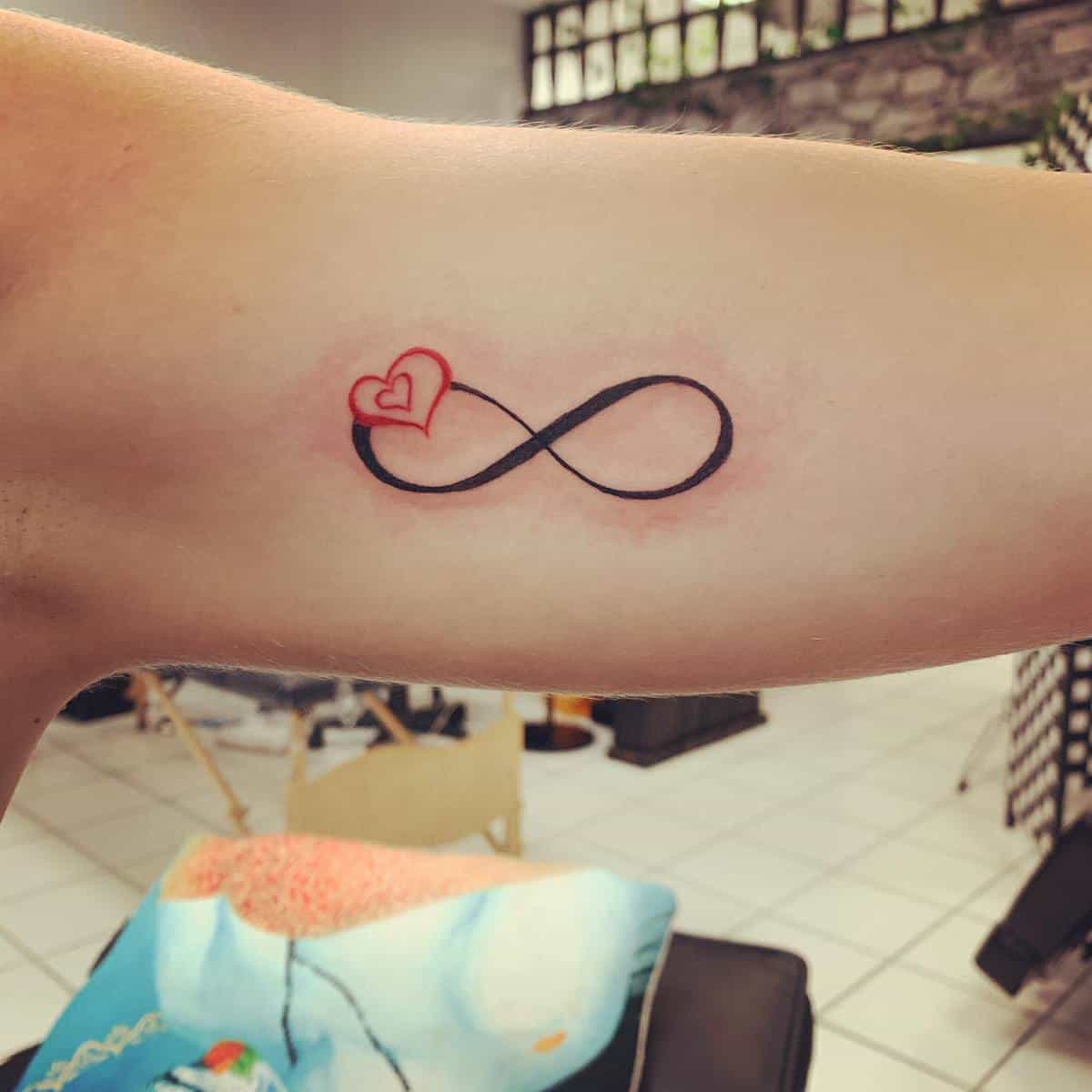 A tattoo of a black infinity symbol with a red heart incorporated into its top left corner on an arm