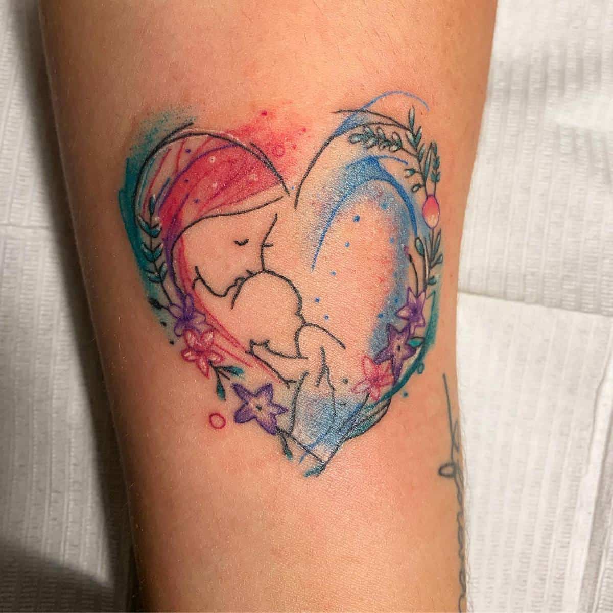 A colorful tattoo featuring a mother holding a baby, surrounded by a heart-shaped floral design